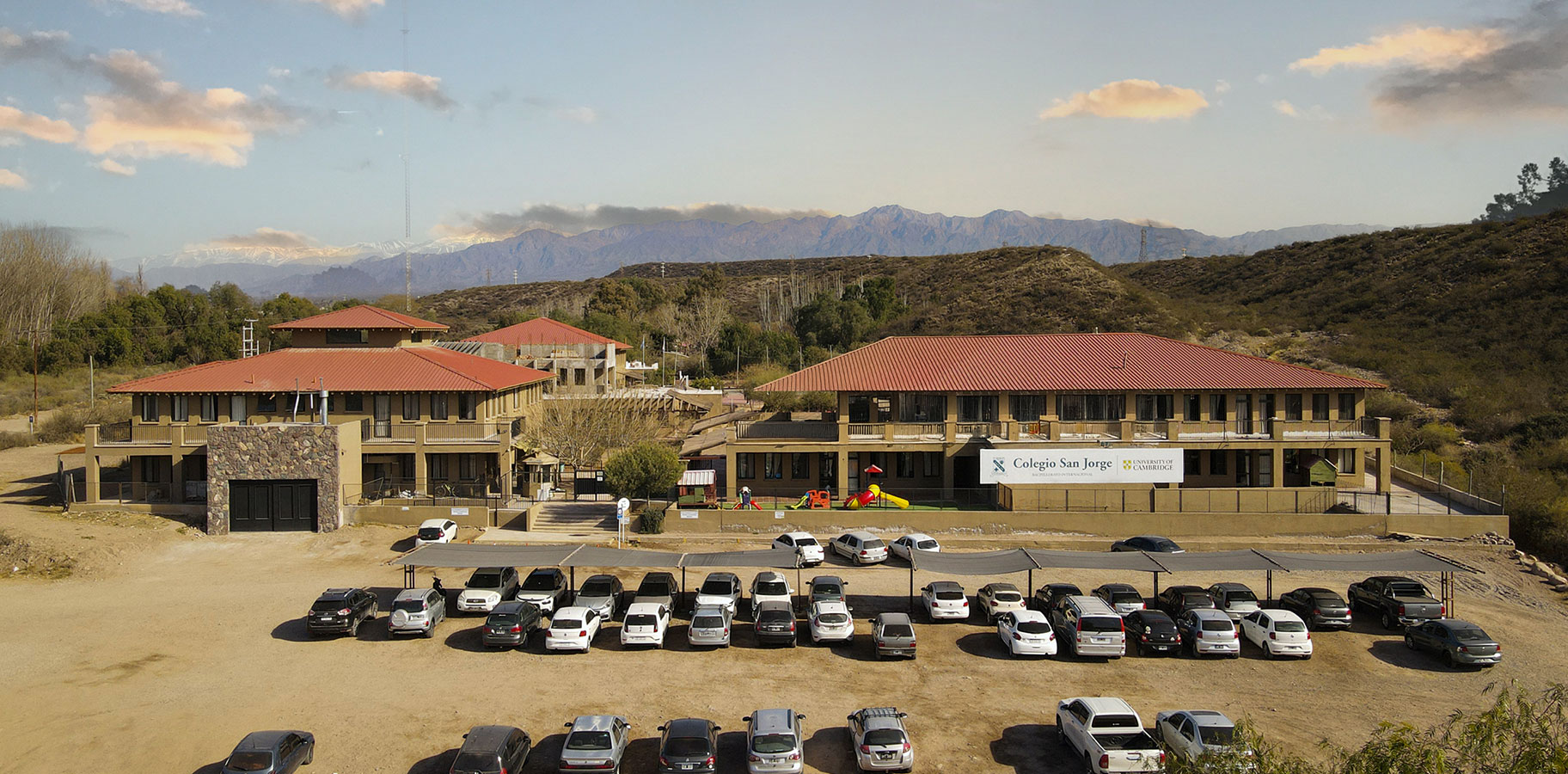 Vista panorámica del Colegio San Jorge con dos edificios principales y un estacionamiento lleno de autos, con montañas al fondo.